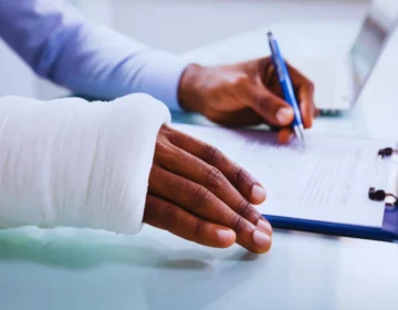 A close-up of a person's hand and forearm wrapped in a white cast or bandage. The person is holding a pen and appears to be writing or filling out a form on a clipboard. Tri-State Work Injuries