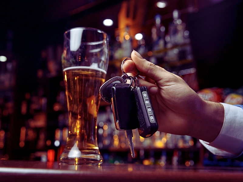 a hand holding car keys next to a glass of beer on a bar counter.