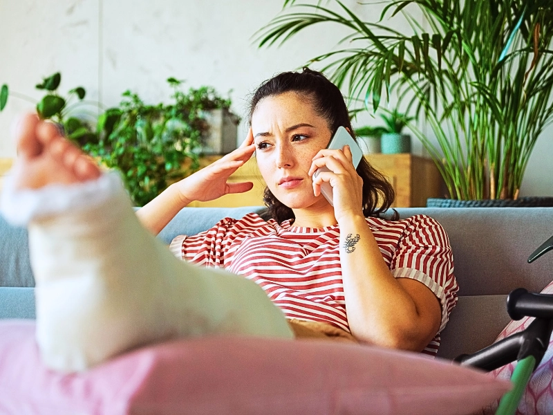 Woman with leg cast talking on smartphone. why insurance keeps delaying your claim