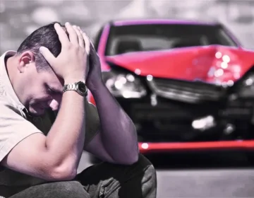 A man is sitting on the sidewalk, clutching his head, completely worried, and in the background, his red car is wrecked from a crash. recover compensation if you’re partially at fault