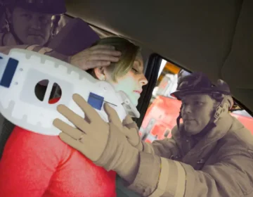 A woman inside a car has her neck immobilized with a neck brace, and a couple of firefighters are assisting her to keep her neck still because she has just been in a car accident. injured in a car accident while working