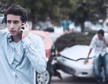 Two boys have been in a car accident and are both on cell phone calls. One of the boys looks noticeably worried. other driver lies about the accident