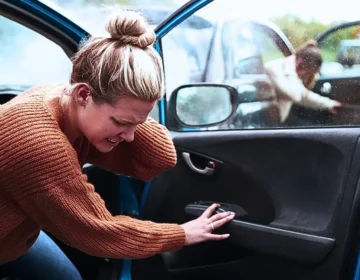 A female passenger getting out of the car is visibly in pain and clutches the back of her neck, having been in an accident that involved a collision with the vehicle in front. Another person is also emerging from the car, injured in the impact. passenger injuries