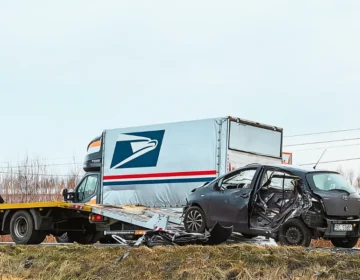 A heavily damaged grey car sits beside a flatbed tow truck on a roadside, with a USPS delivery truck behind them.