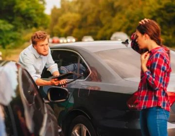 A man looks in a confrontational and annoyed manner at a woman who is clutching her head in worry, as her car has collided with his. car accident involves drivers from different states